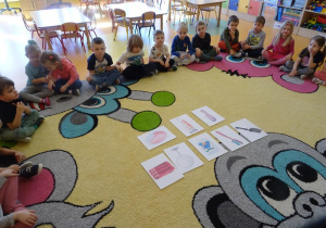 Grupa dzieci siedzi wokół rozłożonych na dywanie ilustracji przedstawiających przedmioty służące do dbania o higienę.